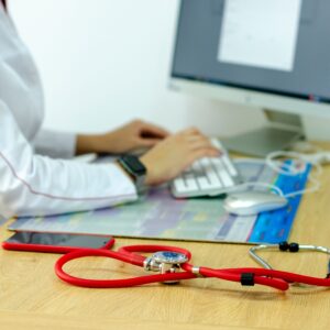 Healthcare professional typing at a desk with a computer, stethoscope on the table and a smartphone nearby, in a blurred office scene.