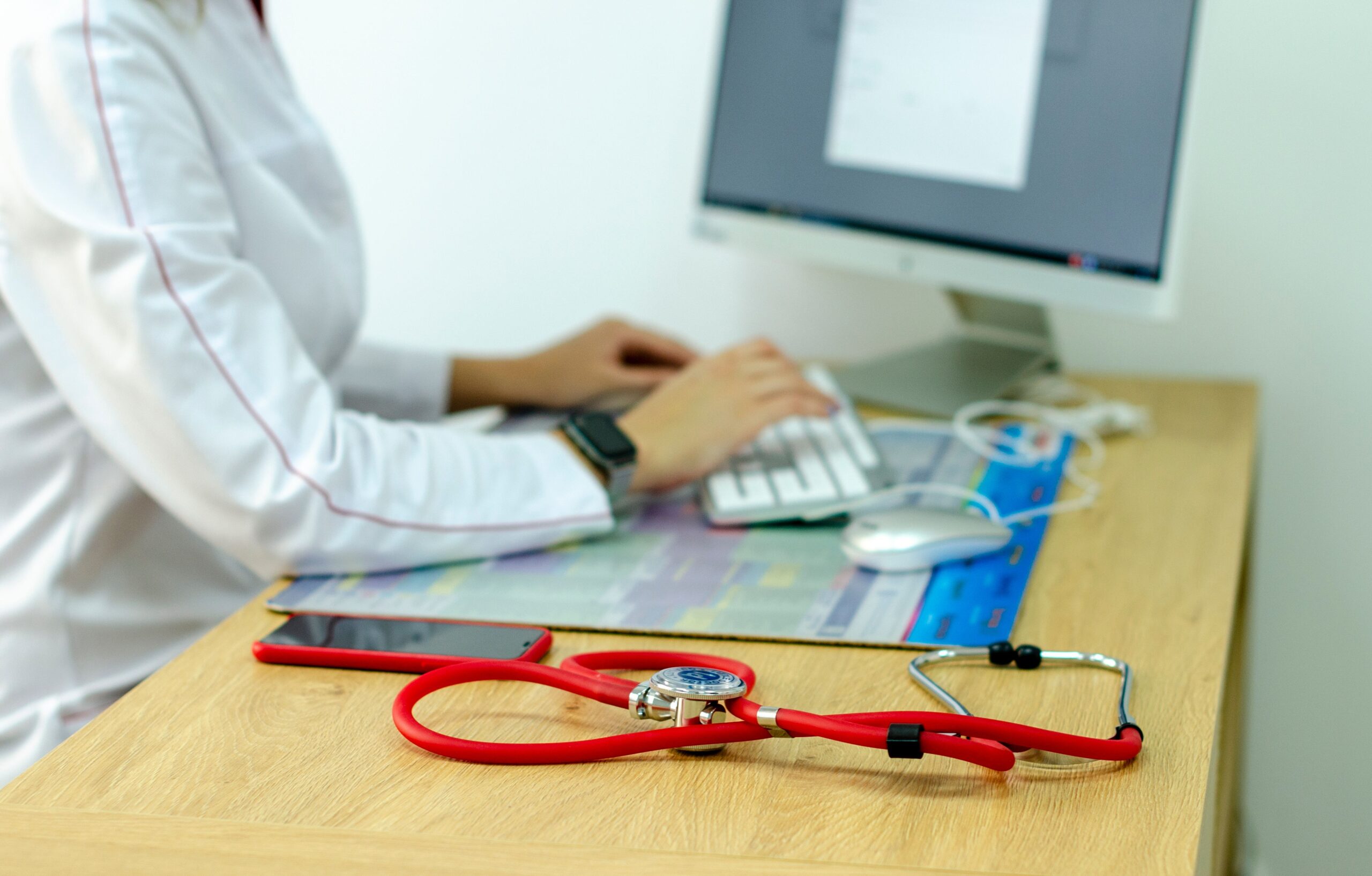 Healthcare professional typing at a desk with a computer, stethoscope on the table and a smartphone nearby, in a blurred office scene.